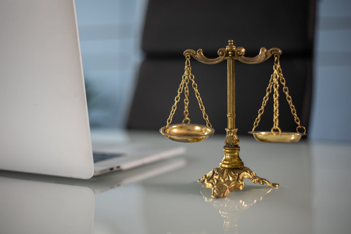 A brass balance scale placed on a reflective office desk next to an open silver laptop, with a black office chair in the background.