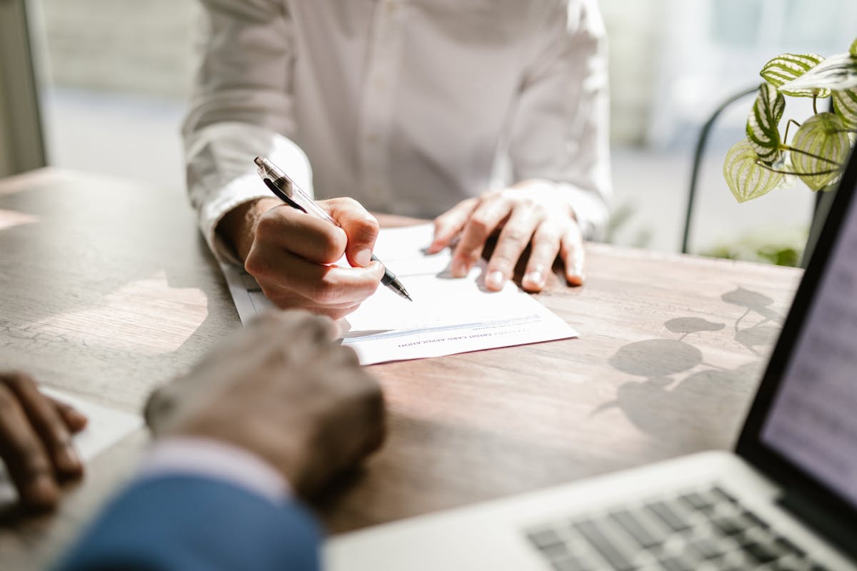 Business person signing a contract at a wooden desk with a laptop nearby. Another professional in a suit is present, indicating a legal or financial agreement in a professional setting.