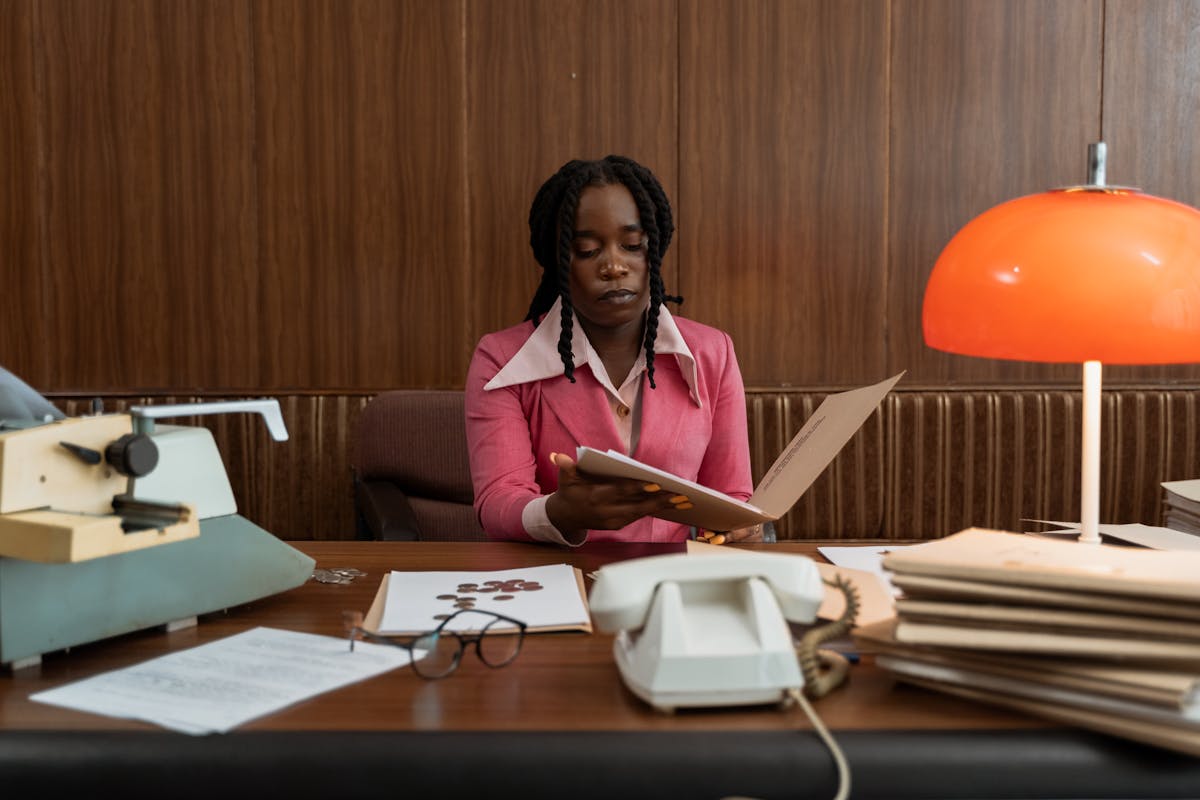 Office scene with a professional woman in a vintage pink blouse reviewing documents at a wooden desk. The workspace features a phone, typewriter, and an orange desk lamp.