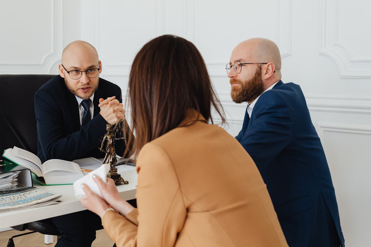 Professional legal consultation in a modern office. Two male lawyers in suits discuss a case with a female client in a beige blazer, with legal books, documents, and a justice scale on the desk.